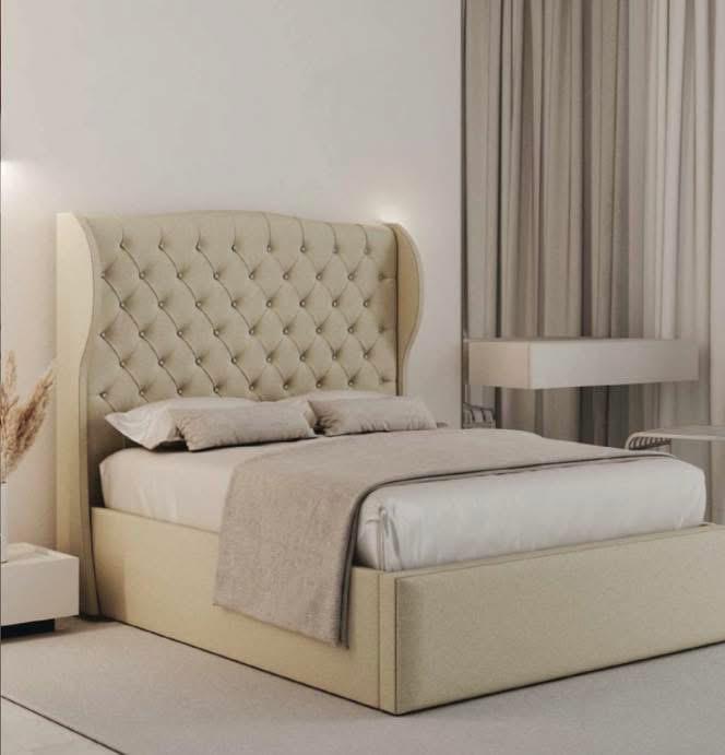 A beige upholstered bed with a tall tufted wingback headboard, shown in a softly lit bedroom setting with neutral bedding, curtains, and a small side table.