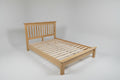 A light natural wood bed frame with slatted base and a simple headboard, shown in a bedroom setting with gray walls, carpet, a window, and a dark dresser.