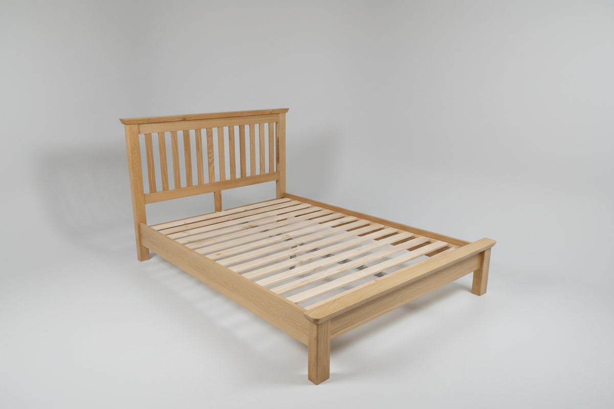 A light natural wood bed frame with slatted base and a simple headboard, shown in a bedroom setting with gray walls, carpet, a window, and a dark dresser.