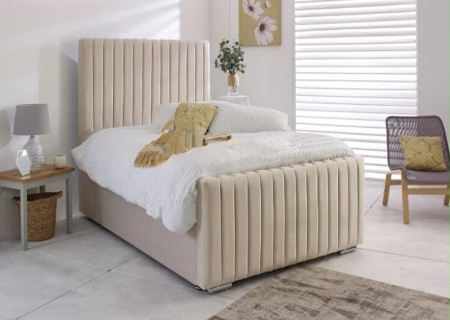 A beige upholstered bed with a tall vertically channel-tufted headboard and matching footboard, styled in a bright bedroom with white bedding, a side table, and a chair.