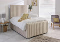 A beige upholstered bed with a tall vertically channel-tufted headboard and matching footboard, styled in a bright bedroom with white bedding, a side table, and a chair.