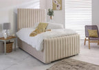A beige upholstered bed with a tall vertically channel-tufted headboard and matching footboard, styled in a bright bedroom with white bedding, a side table, and a chair.
