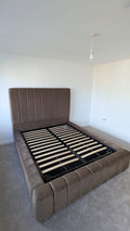 A brown upholstered platform bed frame with a tall padded headboard and matching footboard, shown with the black slatted base exposed in a minimalist bedroom.