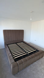 A brown upholstered platform bed frame with a tall padded headboard and matching footboard, shown with the black slatted base exposed in a minimalist bedroom.