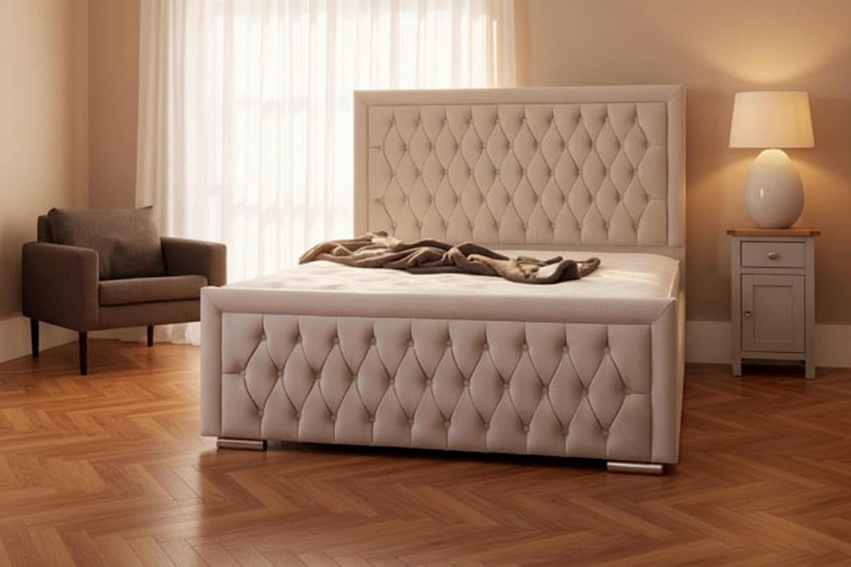 A light gray tufted upholstered bed with a high button-tufted headboard and matching footboard, shown in a bright bedroom setting with a gray armchair, nightstand, lamp, and large windows.