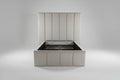 An upholstered platform bed frame with a matching padded headboard in a neutral cream or beige color, shown in a bedroom setting with carpeted floors and a white door in the background.